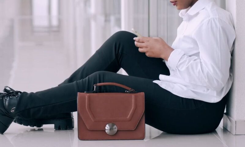 devojka sedi u crnim pantalonama, beloj košulji sa torbicom pored sebe na podu